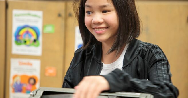 A girl sitting in a classroom using a Perkins Brailler felling what she has written on the paper.
