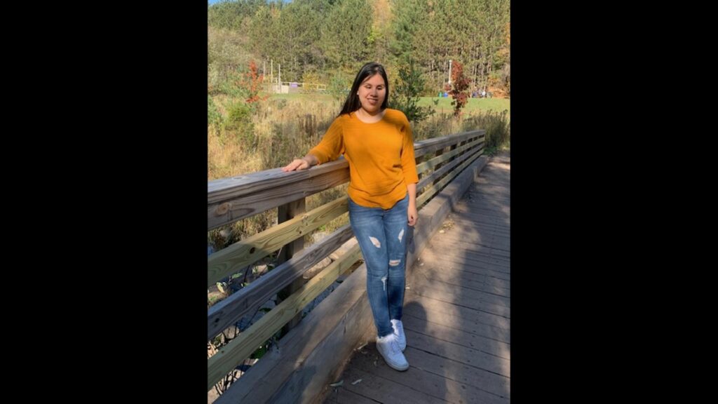 Amy stands on a bridge overlooking a small river. In the background are trees with their leaves starting to change color.